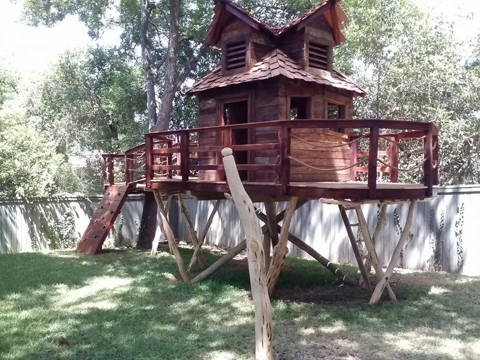 Two-story backyard treehouse on timber stilts with a wraparound deck and climbing wall