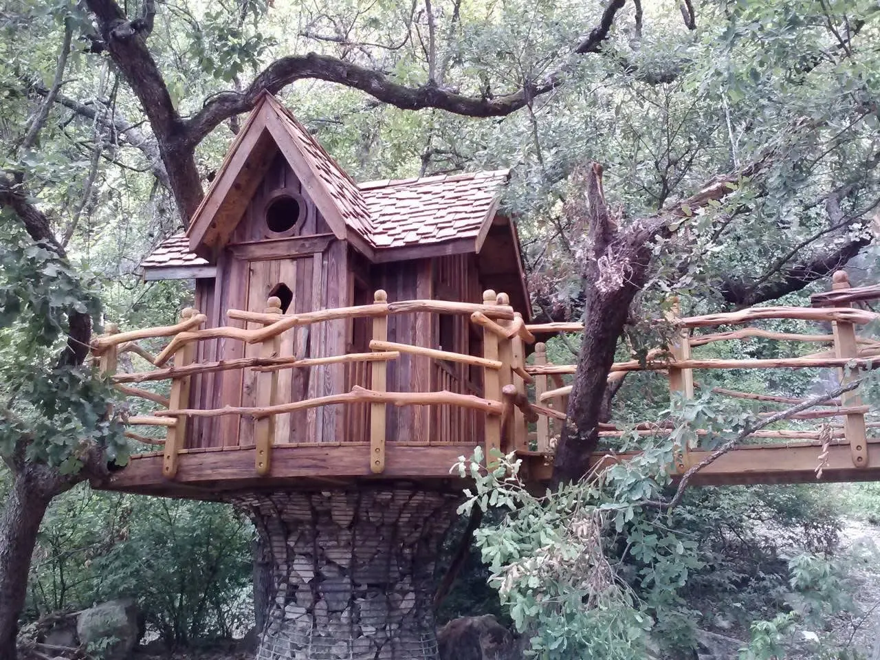 Treehouse with cedar shake roof and rustic branch railings, set on a stone-clad base among oak trees